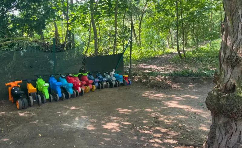 Zahrada klubu pod vzrostlými stromy, u plotu stojí v řadě barevná odrážedla
